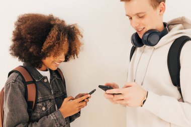 İki üniversite öğrencisi duvarda dikilip akıllı telefonlarına bakıyorlar.