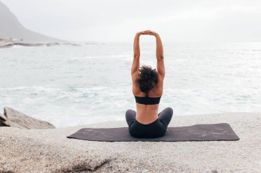 Akşamları okyanus kenarında minderde otururken kollarını geren spor giysili genç bir kadının arka görüntüsü.