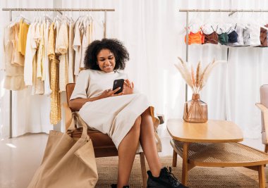 Gülümseyen stil sahibi kadın butikte alışveriş yaparken cep telefonu kullanıyor.