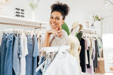 Butikte çalışan gülümseyen bir satıcı. Elinde bir sürü elbise askısı var.