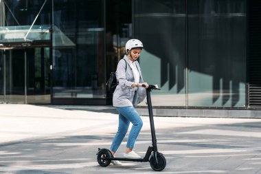 Kafasında kaskı olan genç bir iş kadını ofis binasının yanında elektrikli scooter kullanıyor.