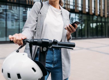 Ofis binasının önünde elektrikli scooterlı ve elinde akıllı bir telefonla duran tanınmamış bir iş kadını. 