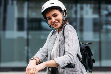 Beyaz bisiklet kasklı neşeli iş kadını portresi. Kameraya bakıyor. Elektrikli scooter gidonuna yaslanıyor.