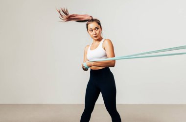 Genç Asyalı sporcu beyaz arka plana karşı direniş bandıyla antrenman yapıyor, başka tarafa bakıyor.