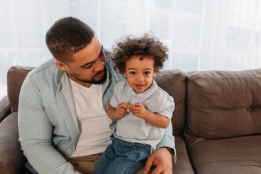 Küçük çocuk babasının kalçasında oturuyor ve kameraya bakıyor. Baba ve oğul kanepede yan yana oturuyor..