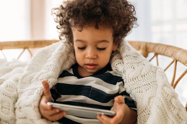 Küçük tatlı bir çocuk, kıvırcık saçlı, koltukta oturmuş, ekosenin altında ve elinde akıllı bir telefon tutuyor.