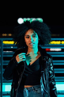 Deri ceketli genç kız neon ışıkların altında poz veriyor.