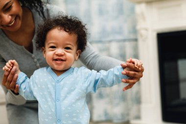 Happy toddler boy learning to walk. Mother assisting son in walking at home.	