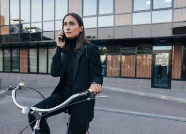 İş kadını dışarıda bisikletle iş binasına karşı otururken bir telefon görüşmesi yapıyor. Siyah iş elbiseli bir kadın cep telefonuyla konuşuyor..