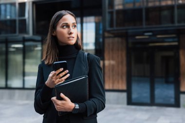 Kendine güvenen ve resmi giyinen ciddi bir kadın cam binanın önünde elinde akıllı telefon ve dijital tablet tutuyor. Siyah giyinmiş genç bir iş kadını dışarıda bekliyor..