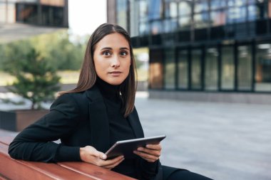 Dijital tabletli genç esmer. Avluda oturan iş kadını siyah resmi kıyafetler giyiyor..