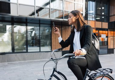 İş kadını bisiklette oturuyor ve cep telefonunu kontrol ediyor. Bisikletin üzerinde otururken cep telefonu kullanan resmi kıyafetli genç bir kadın..