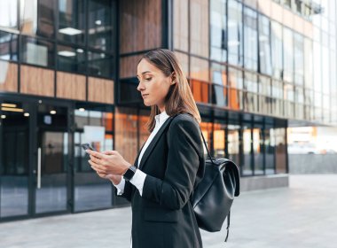 Bir ofis binasının karşısında duran siyah resmi elbiseli, sırt çantalı, cep telefonunda yazan bir kadın.