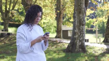 Beyaz kazaklı genç esmer kız cep telefonuyla yazı yazıyor ve doğal yeşil bir ortamda gülümsüyor. Yüksek kalite 4k görüntü