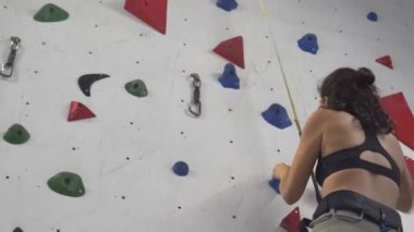 Esmer kız, yapay bir iç mekan duvarına tırmanmayı öğreniyor. Kendini bağlayan bir iple. Spor güvenliği ve sağlıklı yaşam tarzı. Yüksek kalite 4k görüntü