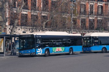 Madrid, İspanya. 15 Şubat 2025. Şehir otobüs durağında mavi bir EMT Madrid otobüsü ve arka planda tarihi binalar.