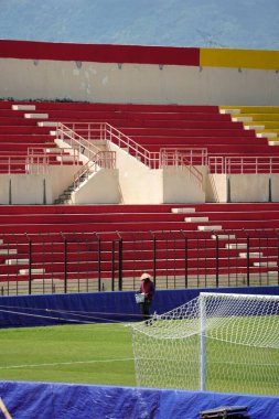 Bir bahçıvan, renkli seyirci koltuklarıyla süslenmiş canlı bir futbol stadyumundaki yemyeşil çimlerle ilgilenir.