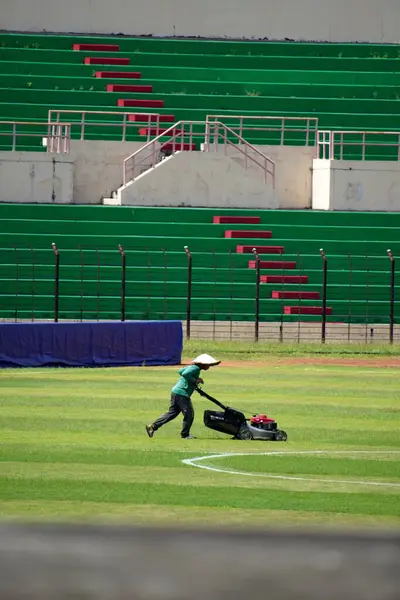Bir bahçıvan, renkli seyirci koltuklarıyla süslenmiş canlı bir futbol stadyumundaki yemyeşil çimlerle ilgilenir.