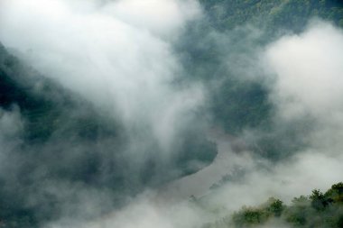 Çikolata-kahverengi suyu olan bir nehir, iki yemyeşil tepeyle çevrili sık bir orman vadisinde esiyor. Sahne, sabah sisi ile örtülmüş ve doğaya mistik bir hava katıyor..