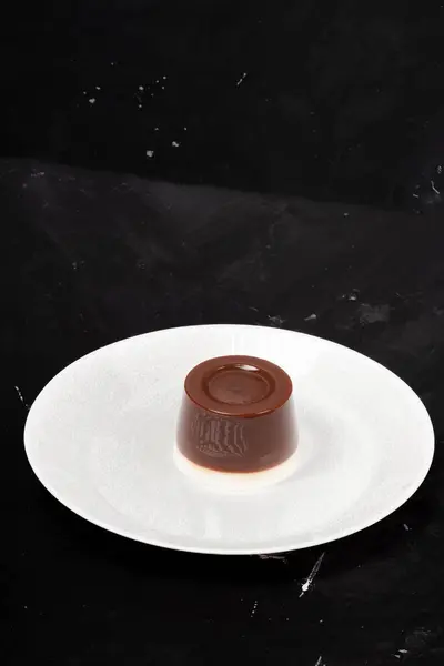 Stock image A glossy chocolate pudding served on a wide white plate, photographed against a dark background, perfect for a dessert treat.