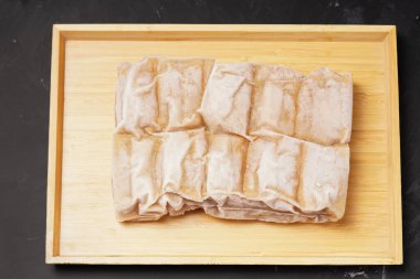 A vacuum-sealed plastic bag containing several frozen tofu bakso, square-shaped tofu with meatball filling tucked in the center, photographed on a wooden tray against a dark background. clipart