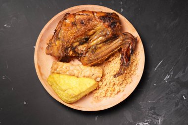 İçinde koyu kahverengi renkte ızgara tavuk, kızarmış tofu ve tempeh ve ahşap bir tabakta sunulan gevrek kırıntılar da var..