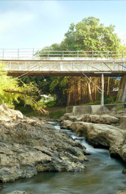 Manzaralı bir nehir lav tortusu kayalarının arasından akar, ağaçlar kıyıları kaplar ve üzerinde uzanan bir köprü vardır. Su kayalık boşluklardan geçip doğal ve huzurlu bir manzara yaratıyor..