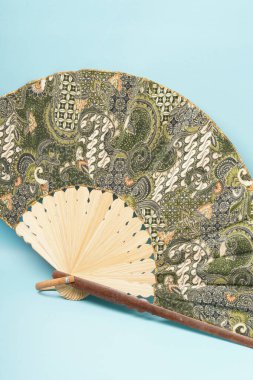 Katlanabilir bambudan yapılmış yeşil batik kumaşla süslenmiş geleneksel bir yarım daire el yelpazesi. Açık mavi bir arkaplanda fotoğraflandı.
