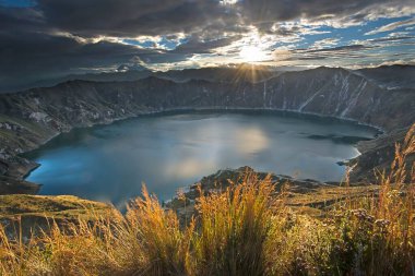 Dramatic sunrise shot of the volcanic caldera and Quilotoa lake and crater. The sun rises over the caldera and reflects on the calm dark water