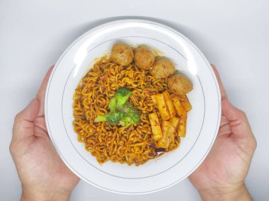 Elinde, üzerinde topoki, köfte ve brokoli olan baharatlı erişte tabağı var.