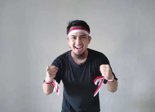 一个年轻的亚洲男人 满面春风 紧紧紧抓住拳头 身穿黑色T恤 头戴旗带 印度尼西亚独立日的概念 — 图库照片