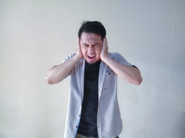 Yetişkin Asyalı adam beyaz gömlek giyip kulaklarını bağıran ifadeyle kapatıyor.