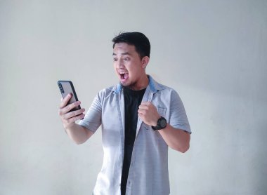 Olgun Asyalı adam cep telefonu tutuyor, yumruğunu sıkıyor ve mutlu bir yüz ifadesi sergiliyor.