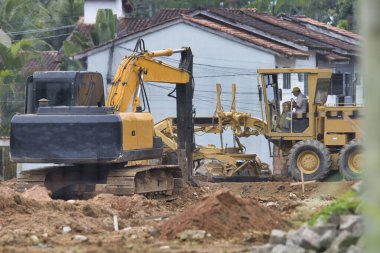 Sarı ve siyah makineli bir şantiye ve sarı kasklı bir adam. Olay yeri kalabalık ve hareketli.