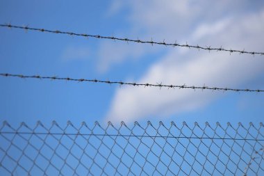 Ön planda tel örgüler var, dikenli tellerle kaplı, bulutlu mavi gökyüzüne karşı.