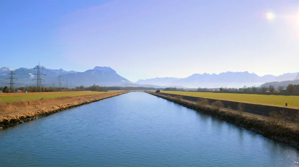 stock image The Rhine River on a summer day between Austria and Switzerland with bicyclist on the dam path and power poles and mountains in the background