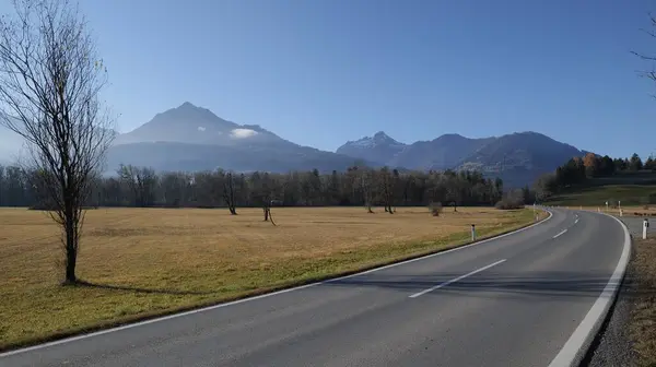 stock image Highway in the sun with curve with large barren field, trees without leaves, mist and mountains in the background