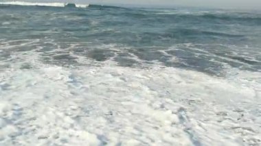 Kocaman dalgaları olan uçsuz bucaksız bir okyanus. Dalgalar kumlu bir sahile çarpıyor ve beyaz köpük oluşturuyor. Gökyüzü beyaz bulutlarla mavidir. Uzakta, sisle kaplı dağlar ve tepeler var..