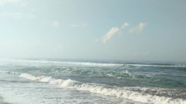 Kocaman dalgaları olan uçsuz bucaksız bir okyanus. Dalgalar kumlu bir sahile çarpıyor ve beyaz köpük oluşturuyor. Gökyüzü beyaz bulutlarla mavidir. Uzakta, sisle kaplı dağlar ve tepeler var..