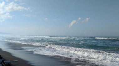 Kocaman dalgaları olan uçsuz bucaksız bir okyanus. Dalgalar kumlu bir sahile çarpıyor ve beyaz köpük oluşturuyor. Gökyüzü beyaz bulutlarla mavidir. Uzakta, sisle kaplı dağlar ve tepeler var..