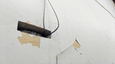 Dikdörtgen delikli bir duvarın yakından görüntüsü. Delik betonla dolu ve duvar boyunca uzanan elektrik kabloları var. Duvar beyaza boyanmış ama soyulmuş boya parçaları var..