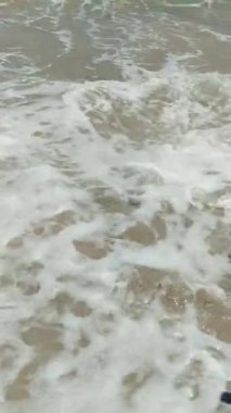 Çalkantılı dalgalar kumlu sahilde köpük ve tortuyu karıştırıyor. Su bulanık, yüzeyin altındaki kum ve kaya parçalarını ortaya çıkarıyor.