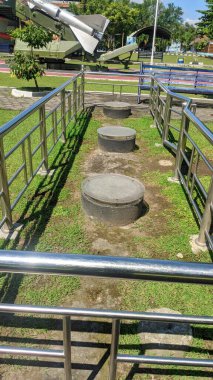 Metal parmaklıklarla çevrili çimenli bir alanda metal kaplamalı üç sıra beton kuyu başı ve arka planda bir füze görüntüsü var..