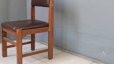 Kahverengi minderli, taşralı ahşap bir sandalye köşede oturur. Arka planda, eski veya minimalist bir iç mekan olduğunu gösteren desenli gri bir duvar ve döşeme var..