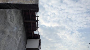 Yıpranmış bir binanın ve çatısının bir kısmının bulutlu bir gökyüzüne bakan solucan bakışı görüntüsü. Koyu renk, desenli duvar ve çatıdaki ahşap kirişler çerçevenin sol tarafından görülebilir.