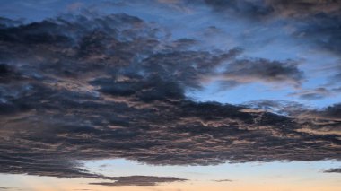 Katmanlı bulutlarla dolu dinamik bir gökyüzü. Koyu, desenli fırtına bulutları, güneşin doğuşundan kalma turuncu ve pembe ipuçları ile vurgulanır. Tepesinde açık, soluk mavi bir gökyüzü vardır.