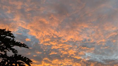 Dinamik bir gökyüzü dokulu bir bulut mozaiğiyle dolu. Karanlık, karamsar gri ve mavi bulutlar güneşin doğuşundan gelen parlak, ateşli portakallarla iç içe geçerek dramatik ve soyut bir desen oluştururlar..