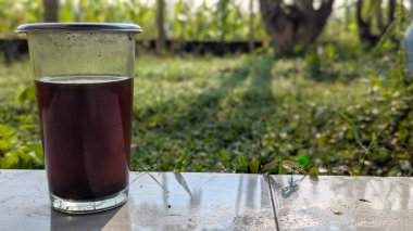 Bir bardak koyu sıvı, muhtemelen kahve, fayanslı bir yüzeyde. Arka plan bulanık yeşil bir çimenlik ve ağaçtır. Sabah güneşi yumuşak bir gölge bırakır..