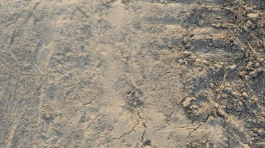 Kurumuş toprak, ince toz ve küçük toprak kütleleri çatlak belirtileri gösteriyor. Daha sert, daha büyük toprak parçaları ve kuru bitki örtüsü sağ tarafta görülebilir. Toprak açık, tozlu kahverengi..