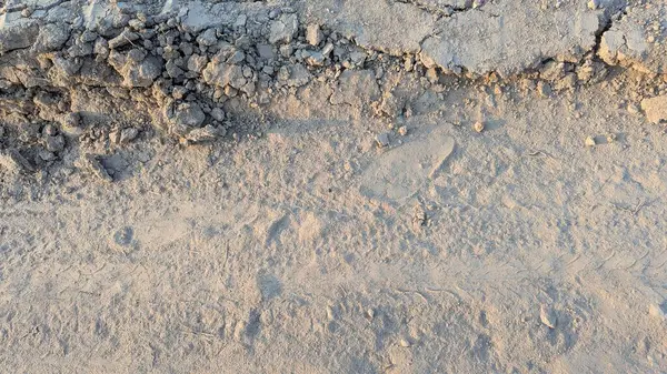 İnce, tozlu zemin ile daha büyük, çatlamış toprak ve kayalar arasında belirgin bir ayrım olan toprak bir yol manzarası. Ayak izleri tozun içinde görünür..